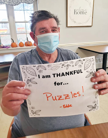 older man sitting in chair holding sign that says I am Thankful for Puzzles
