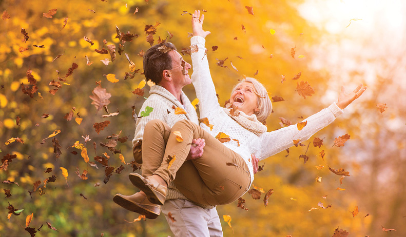 older couple enjoying the fall season. Husband carrying wife through falling leaves.